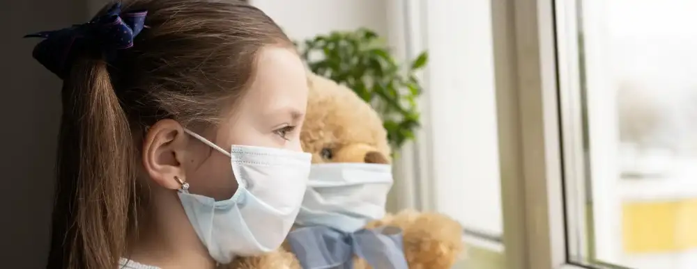 Girl and teddy bear looking out the window. They both have masks on.